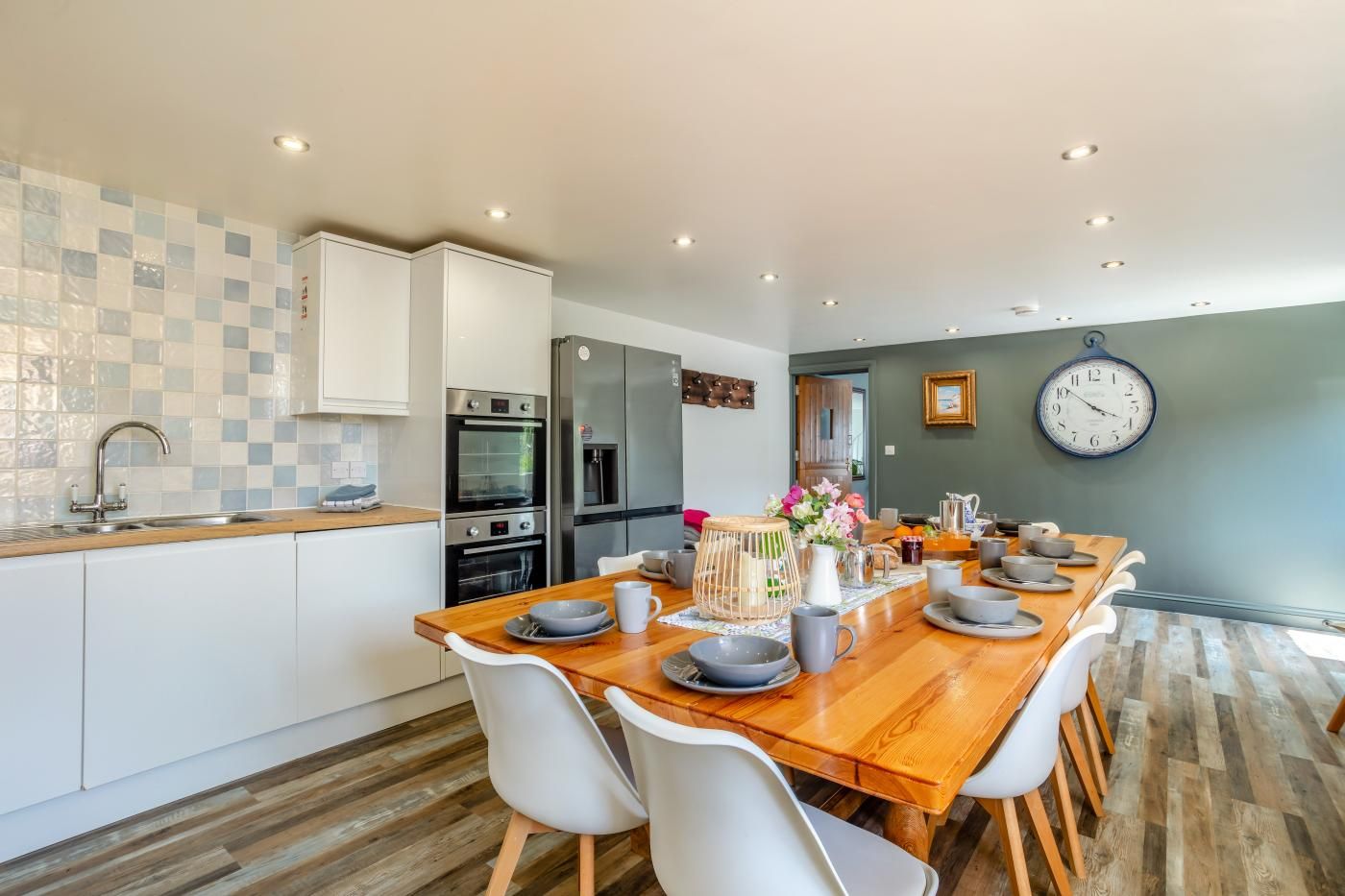 A modern kitchen with white cabinets, a wooden dining table, and a clock on the wall.