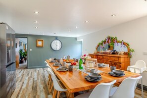 A dining area with a wooden table, white chairs, a clock, and a decorative mirror. - 6 Bed in Holsworthy (oc-89218) (Holsworthy Hamlets)
