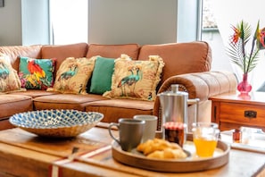 Un canapé en cuir marron agrémenté de coussins colorés, une table d’appoint en bois sur laquelle se trouvent une cafetière, un pichet en verre, un plateau avec des pâtisseries et un verre de jus d’orange.