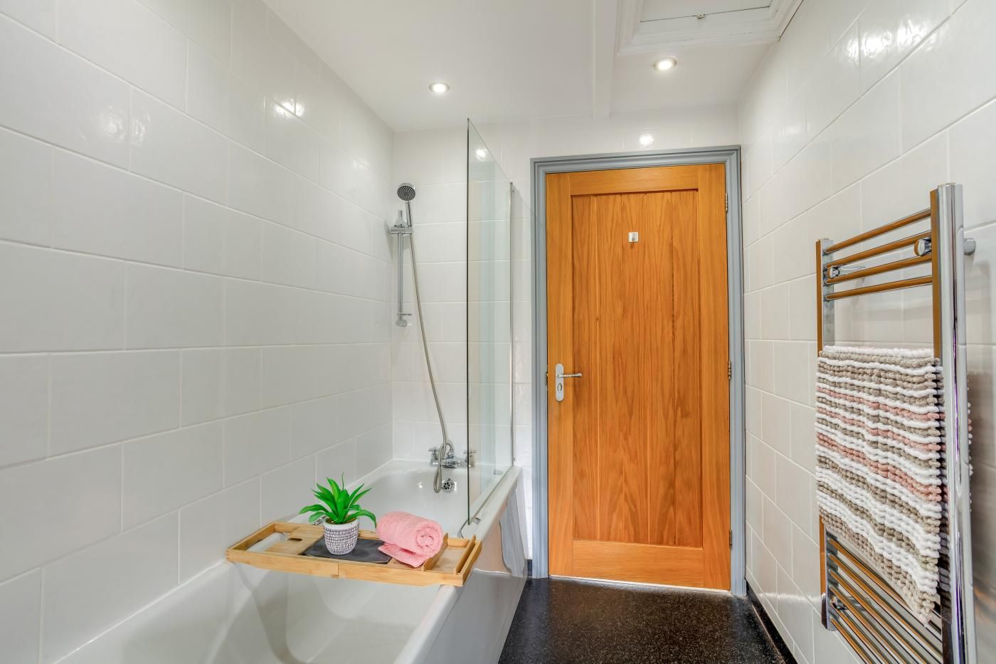 A bathroom with white tiles, a wooden door, a glass shower enclosure, a towel rack, and a small plant on a tray.