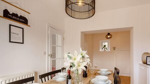 Un comedor con una mesa de madera dispuesta para cuatro, platos blancos, vasos y cubiertos. Hay un florero con flores blancas sobre la mesa y un cartel enmarcado en la pared.