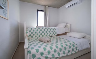 Una cama individual con una colcha estampada, una mesita de noche, una ventana con cortinas y un equipo de aire acondicionado.