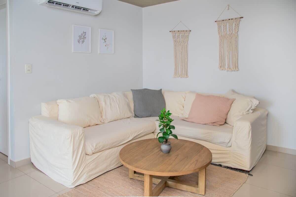 En stue med en beige sofa, et rundt træbord og en potteplante.