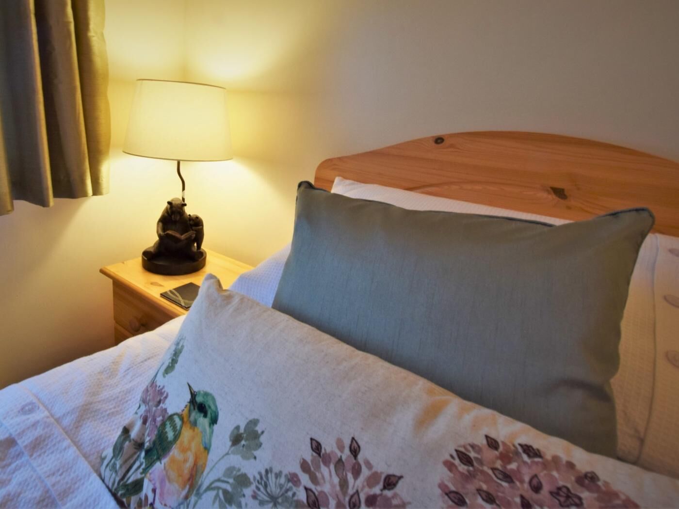 Een net opgemaakt bed met een bloemenkussen, een houten hoofdbord, een bedlampje en een nachtkastje met een klein beeldje.