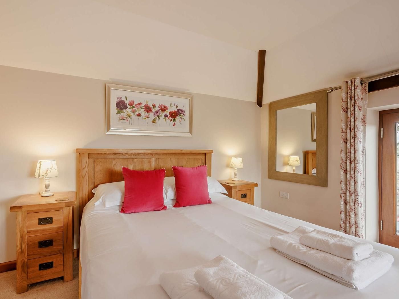 A hotel room with a large bed, two bedside tables with lamps, a floral artwork above the bed, a mirror on the wall, and a door with curtains.