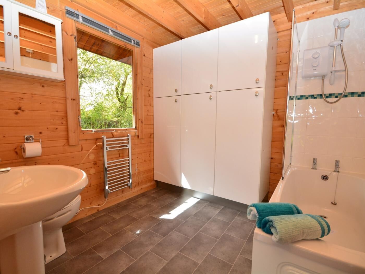 Een houten badkamer in een blokhut met toilet, wastafel, kastjes, een raam en een douche.