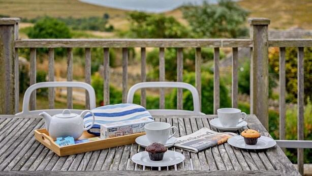 Une terrasse en bois avec une table dressée pour le thé et les biscuits, offrant une vue sur un paysage vallonné.