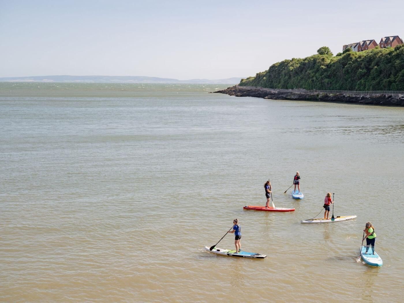 Eine Gruppe von Menschen steht auf Paddleboards auf ruhigem Wasser, im Hintergrund eine felsige Küste und Gebäude.