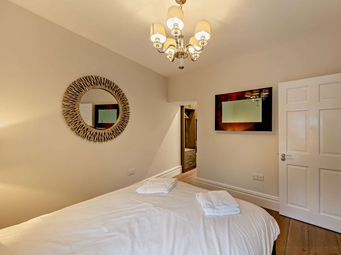 A hotel room with a bed, a round mirror, a chandelier, a door, and a wall-mounted artwork.