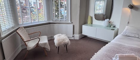 A bedroom with a bed, a chair, a small stool, a vanity with a mirror, and a radiator.
