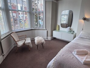 Une chambre à coucher comprenant un lit, une chaise, un petit tabouret, une coiffeuse avec un miroir et un radiateur.