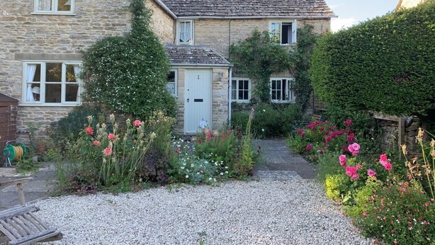 A stone house with a gravel driveway, surrounded by a garden with various flowers and plants. - 2 Bed in Cirencester (oc-hic05) (Eastleach)