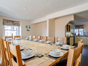 Un comedor con una mesa larga dispuesta para una comida, sillas de madera y vista a la cocina.