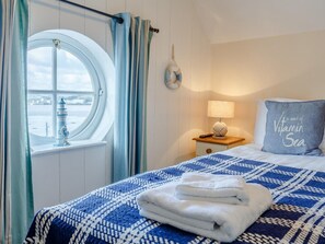 A bedroom with a bed, a nightstand, a lamp, a pillow with a message, a window with curtains, and a view of the sea.