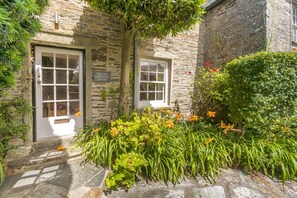 A stone building with two white-framed windows and a door, surrounded by lush greenery and flowering plants. - 4 Bed in Tintagel (oc-tolbo) (Bossiney)