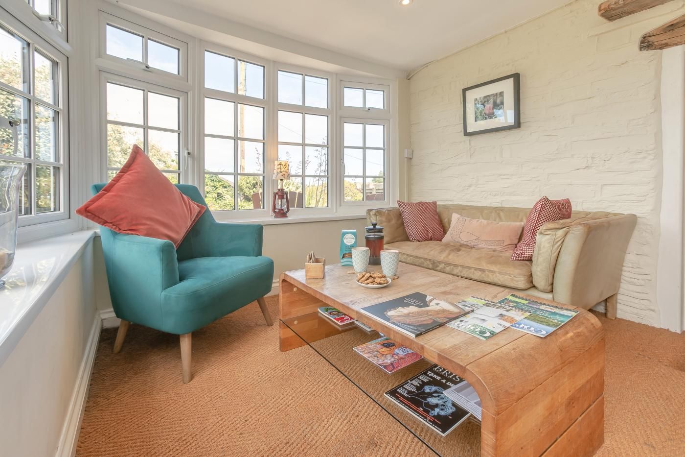 A cozy living room with a teal armchair, a wooden coffee table with magazines, a beige sofa with patterned cushions, and a white brick wall.