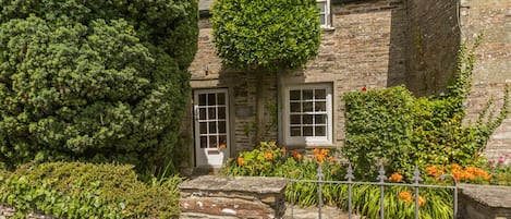 A stone building with a door and windows, surrounded by greenery and a stone pathway.