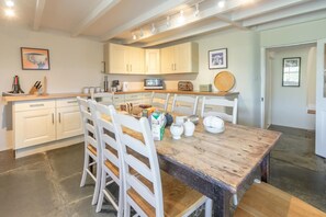 A kitchen with white cabinets, a wooden dining table, and chairs. There are kitchen appliances like a microwave and a kettle. - 4 Bed in Tintagel (oc-tolbo) (Bossiney)