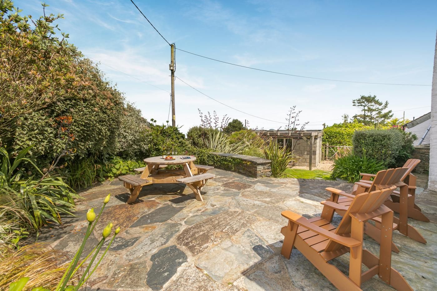 A stone patio with a wooden table and chairs, surrounded by lush greenery and a stone wall.