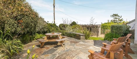 A stone patio with a wooden table and chairs, surrounded by lush greenery and a stone wall.