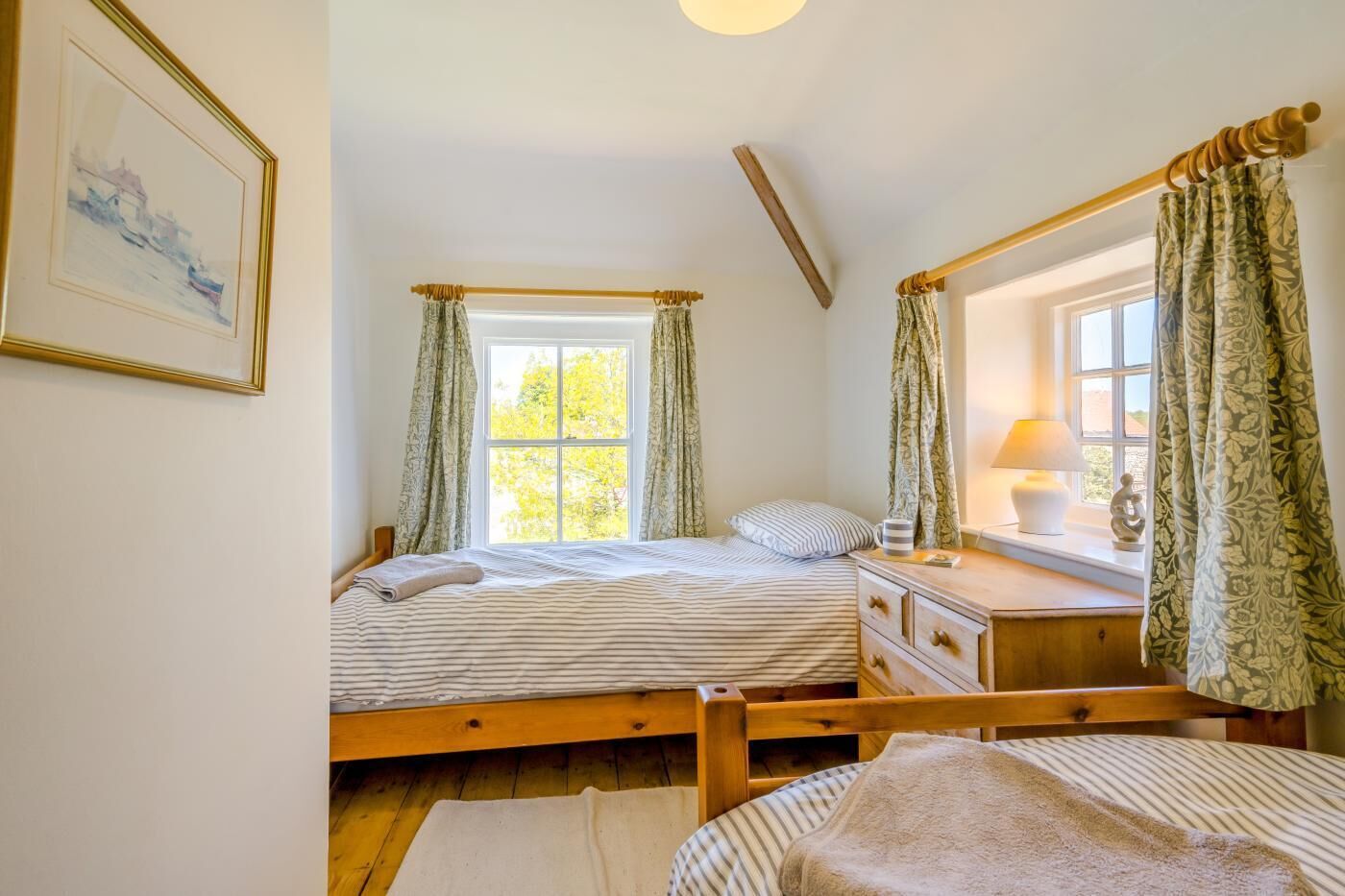 A bedroom with two beds, a bedside table with a lamp, a window with curtains, and a framed picture on the wall.