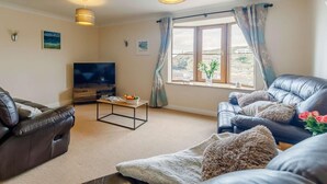 A living room with a TV, leather sofas, a coffee table, and a large window with curtains. - 4 Bed in Redruth (oc-26386) (Portreath)