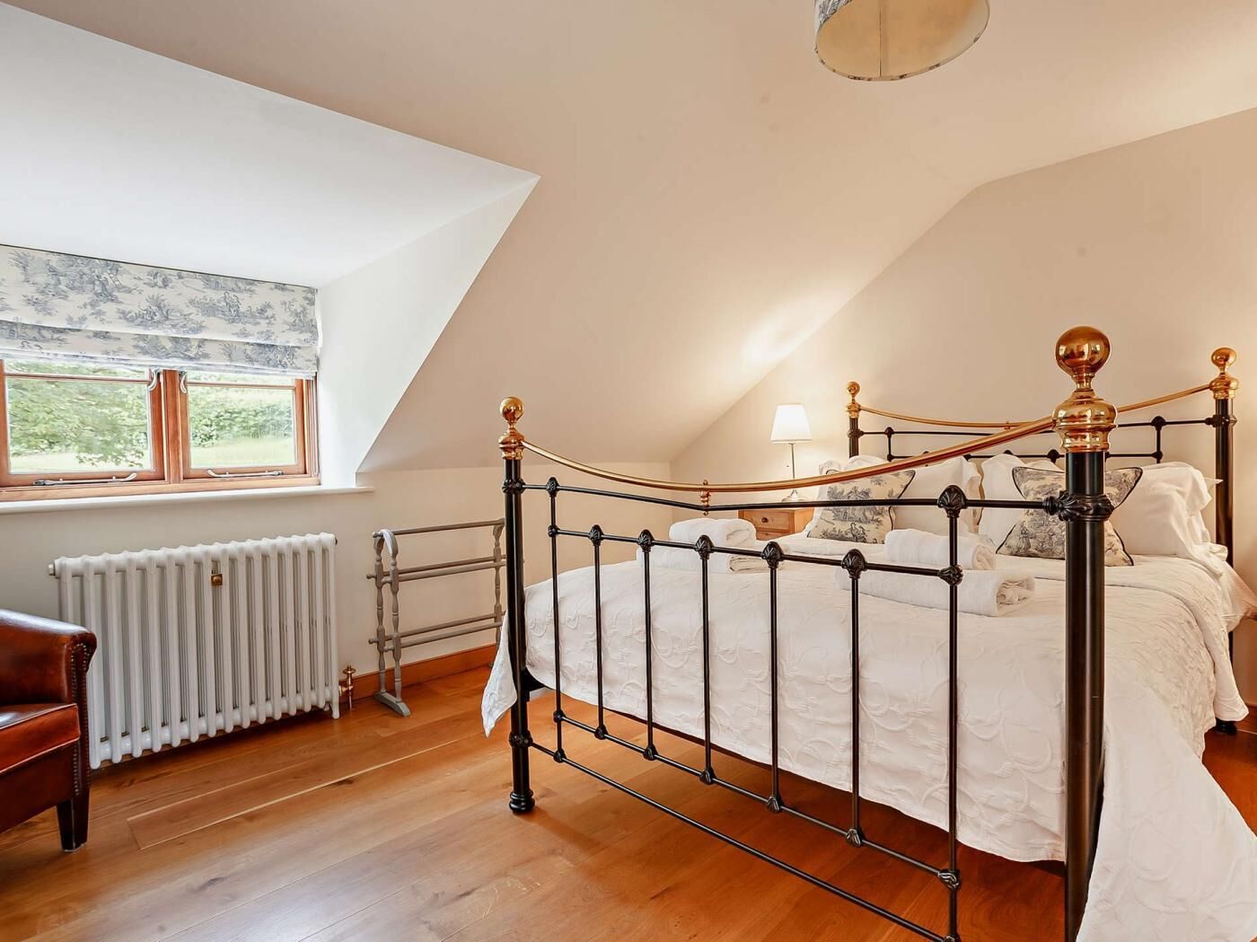 Een slaapkamer met een schuine plafond, een bed met metalen frame, een radiator, een raam met uitzicht en een stoel.