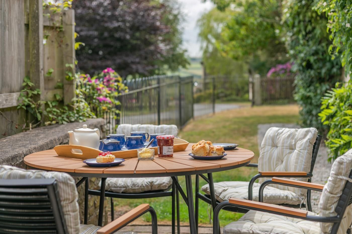 Une table ronde en bois, sur laquelle sont disposés un service à thé, des scones et de la confiture, entourée de chaises rembourrées, le tout dans un cadre de jardin.
