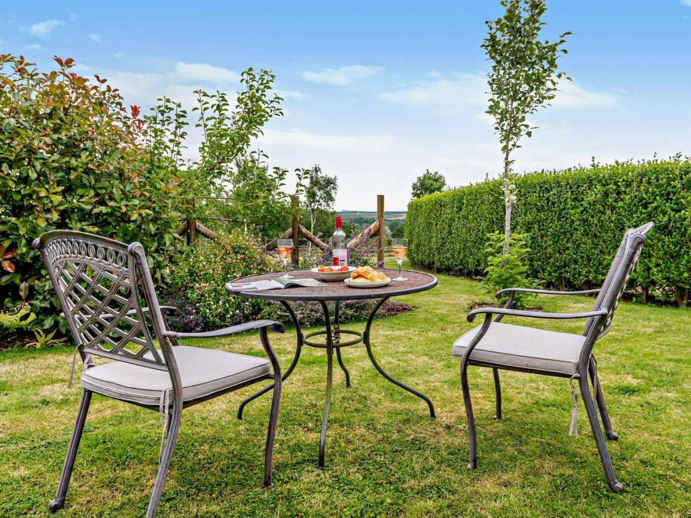 A garden patio with two chairs and a table, surrounded by greenery and a hedge.