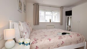 Um quarto com uma cama, uma mesa de cabeceira com abajur e canecas, uma janela com cortinas e um radiador.