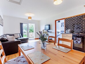 A modern living room with a sofa, a dining table with placemats, and a kitchen area with a stove and a TV. - 2 Bed in Bristol (oc-37147) (Chew Magna)