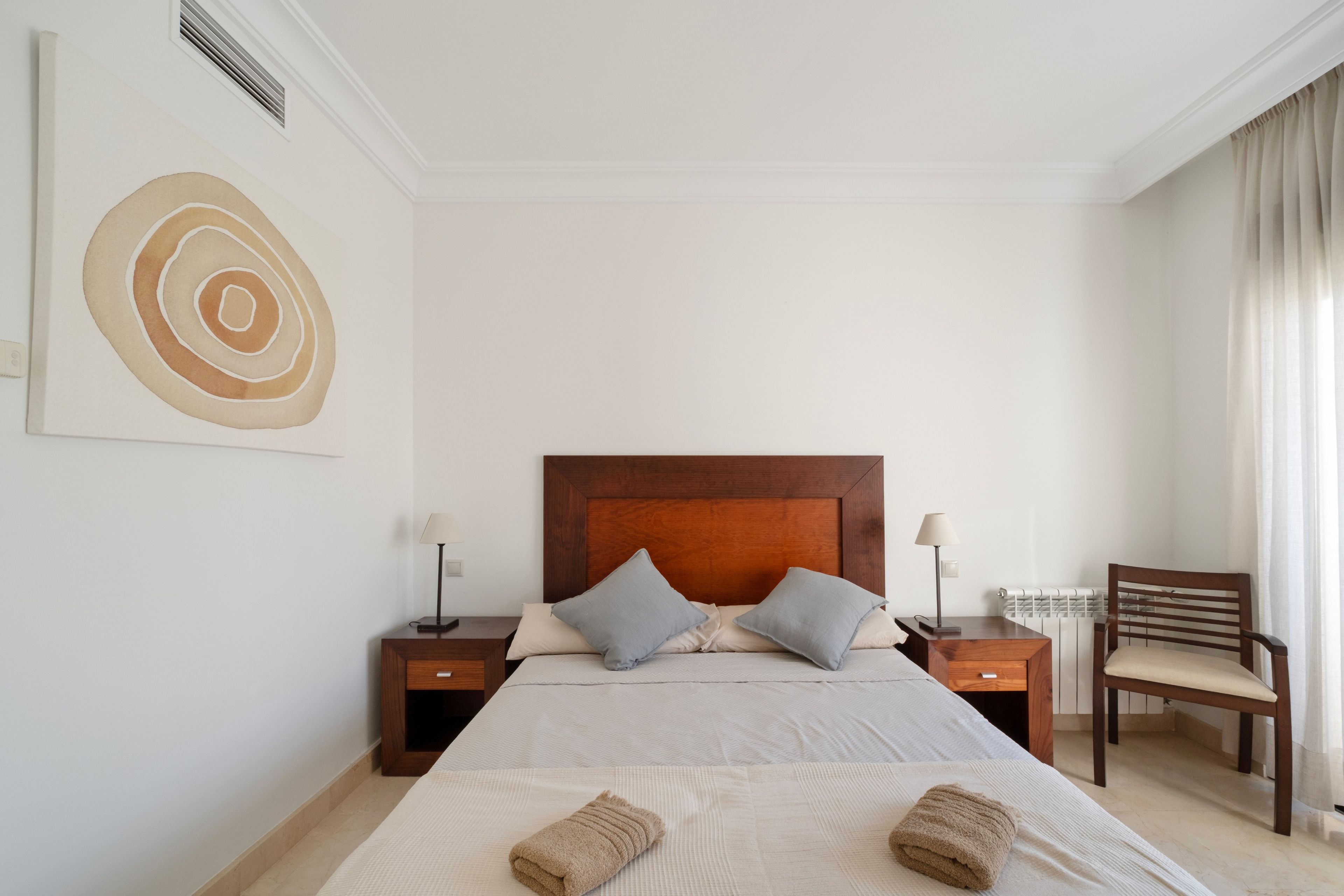 Habitación de hotel ordenada con una cama grande, dos mesitas de noche, una silla y una obra de arte en la pared.