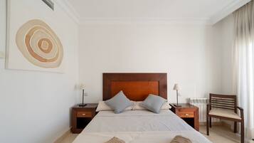 Habitación de hotel ordenada con una cama grande, dos mesitas de noche, una silla y una obra de arte en la pared.
