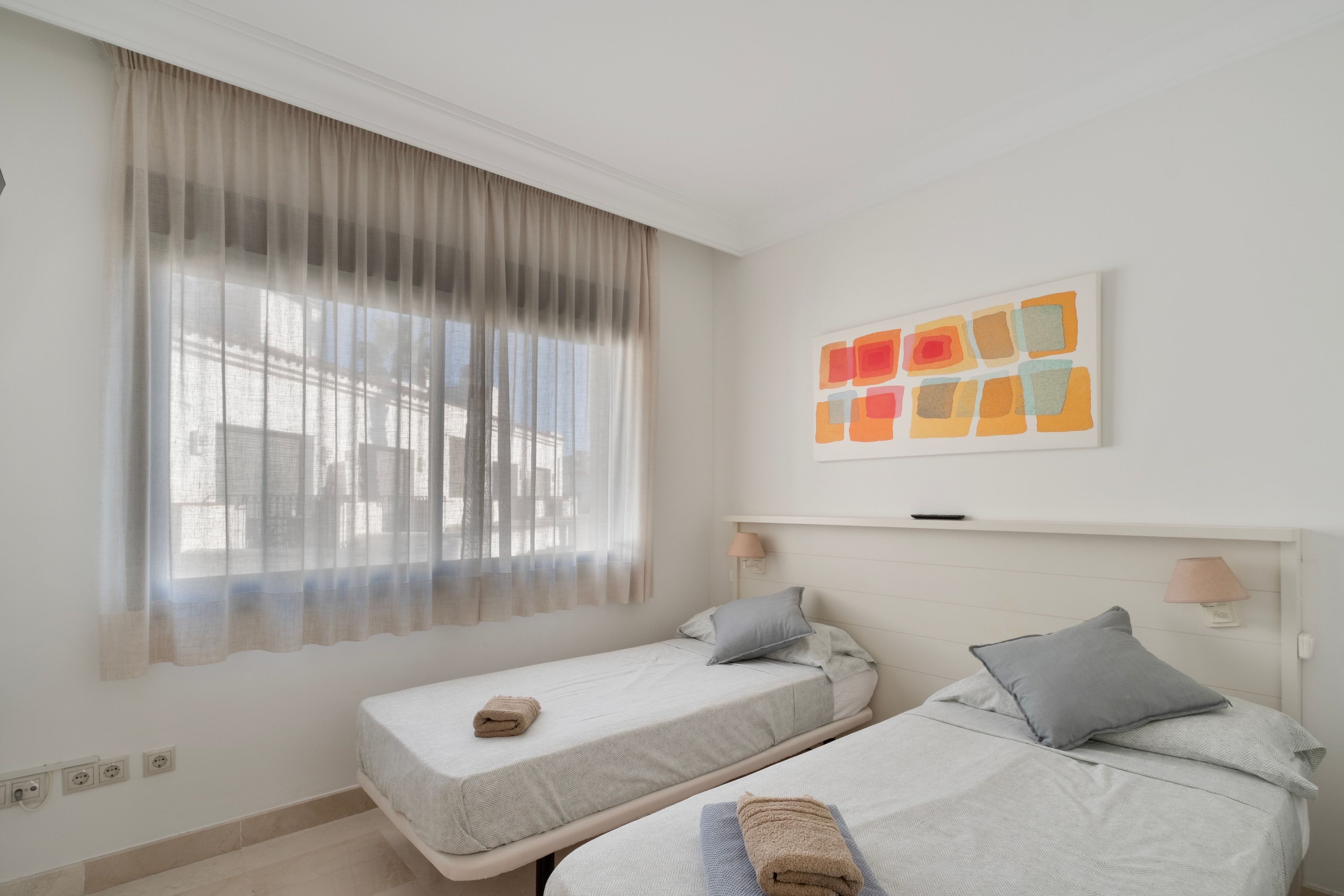 Una habitación con dos camas, una ventana con cortinas translúcidas, una obra de arte en la pared y lámparas de noche.