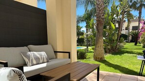 Un patio con sofá y mesa, rodeado de un jardín con palmeras y una piscina.