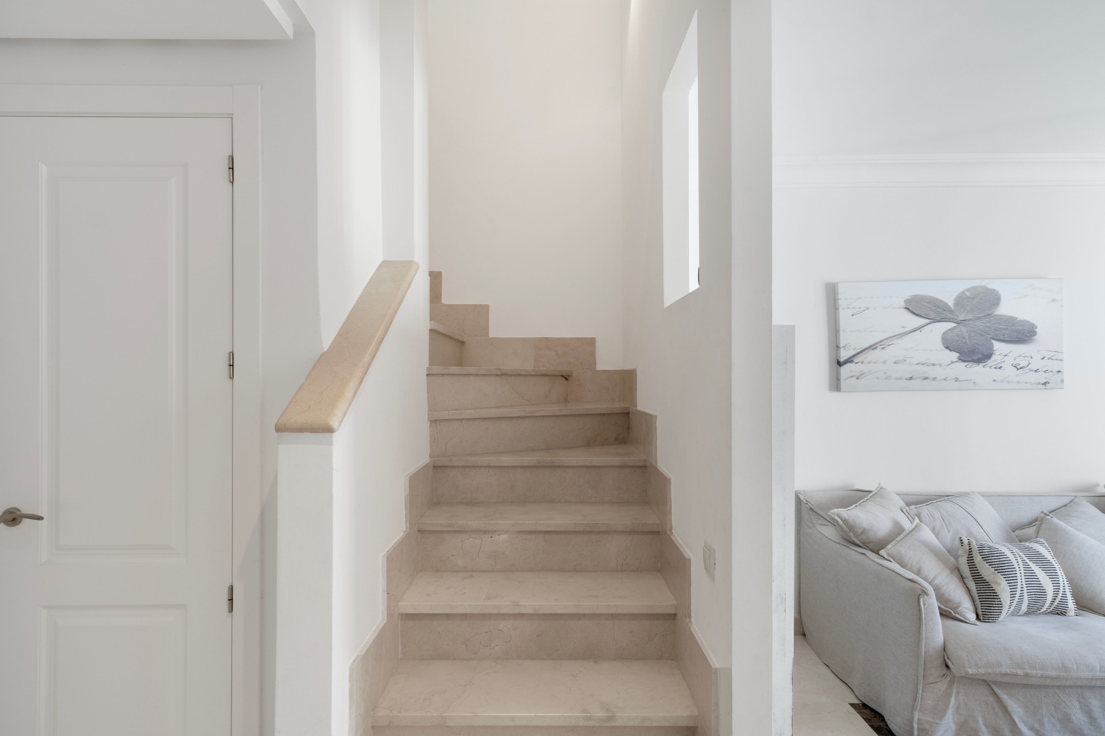 Una escalera con peldaños color beige que lleva a un rellano, una puerta blanca a la izquierda y un sofá gris con cojines estampados a la derecha.