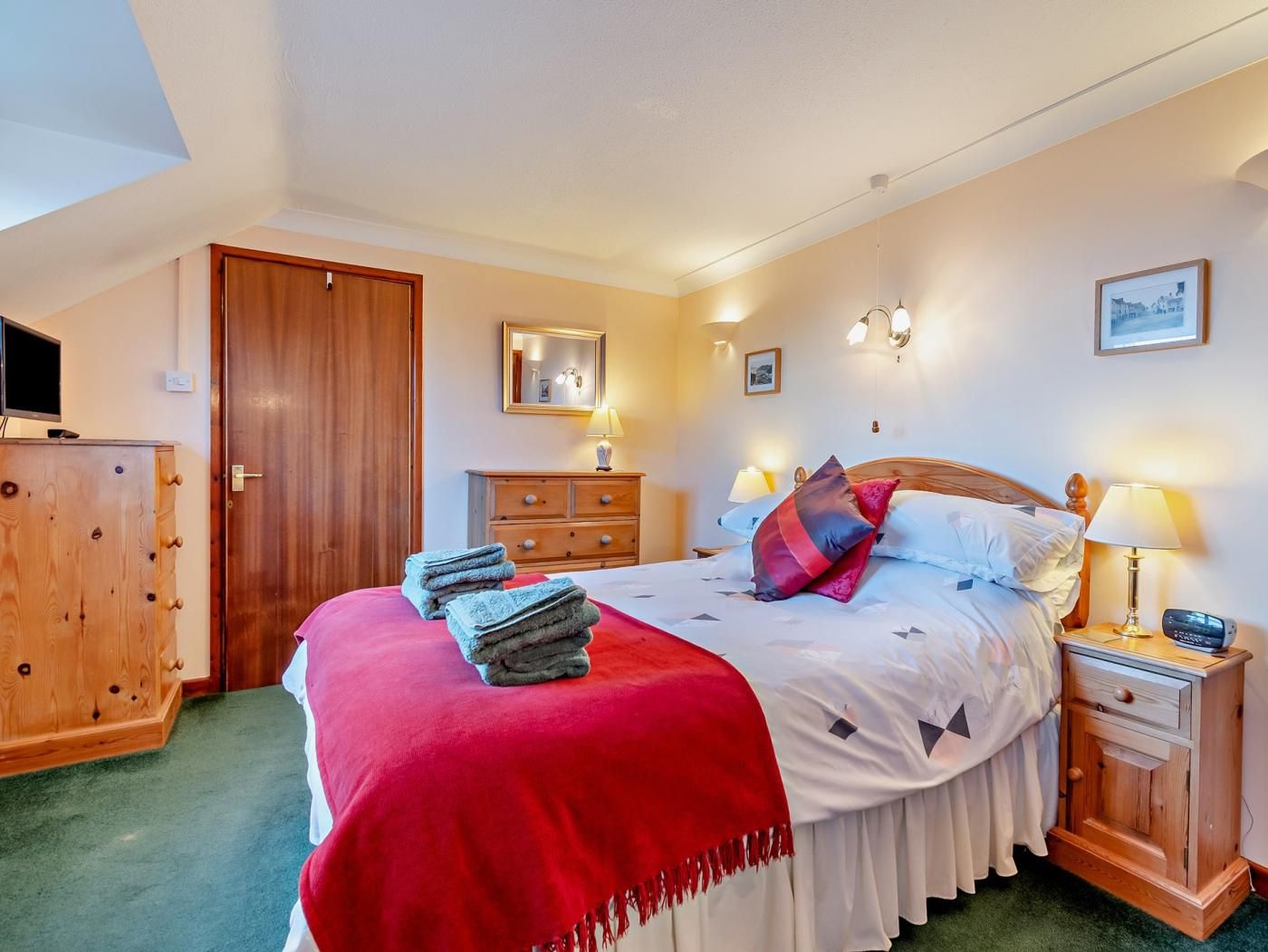 A hotel room with a bed, a wooden wardrobe, a television, a desk with a lamp, and a wall-mounted mirror.