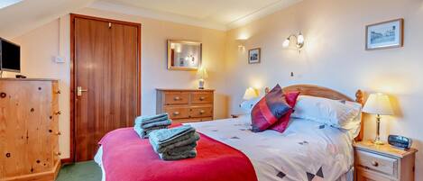 A hotel room with a bed, a wooden wardrobe, a television, a desk with a lamp, and a wall-mounted mirror.