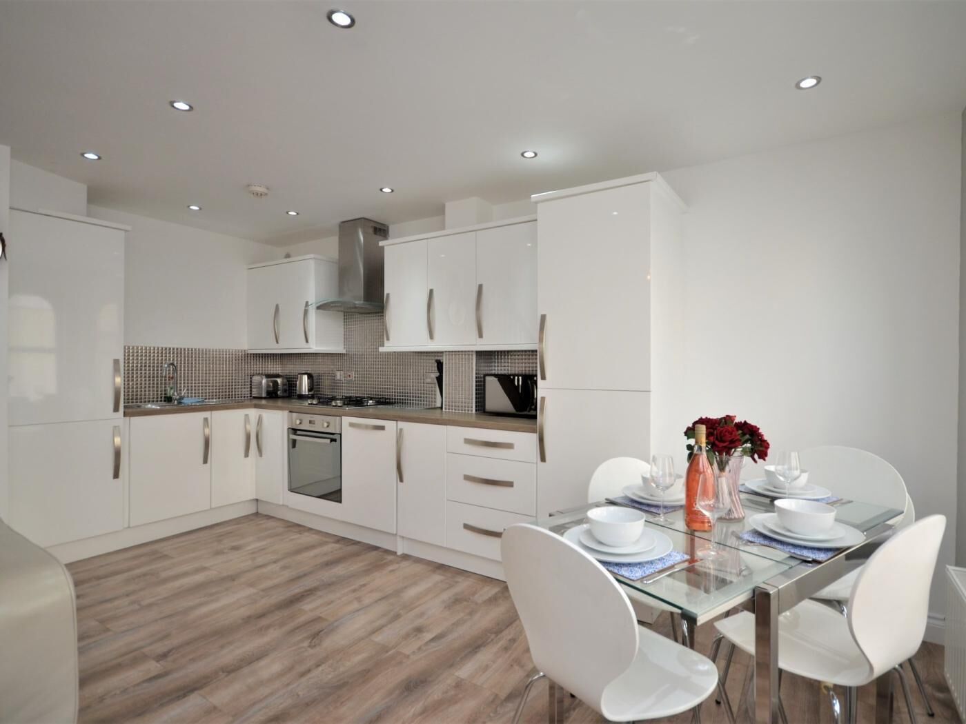 A modern kitchen with white cabinets, a stainless steel range hood, and a dining area with a glass table and white chairs.