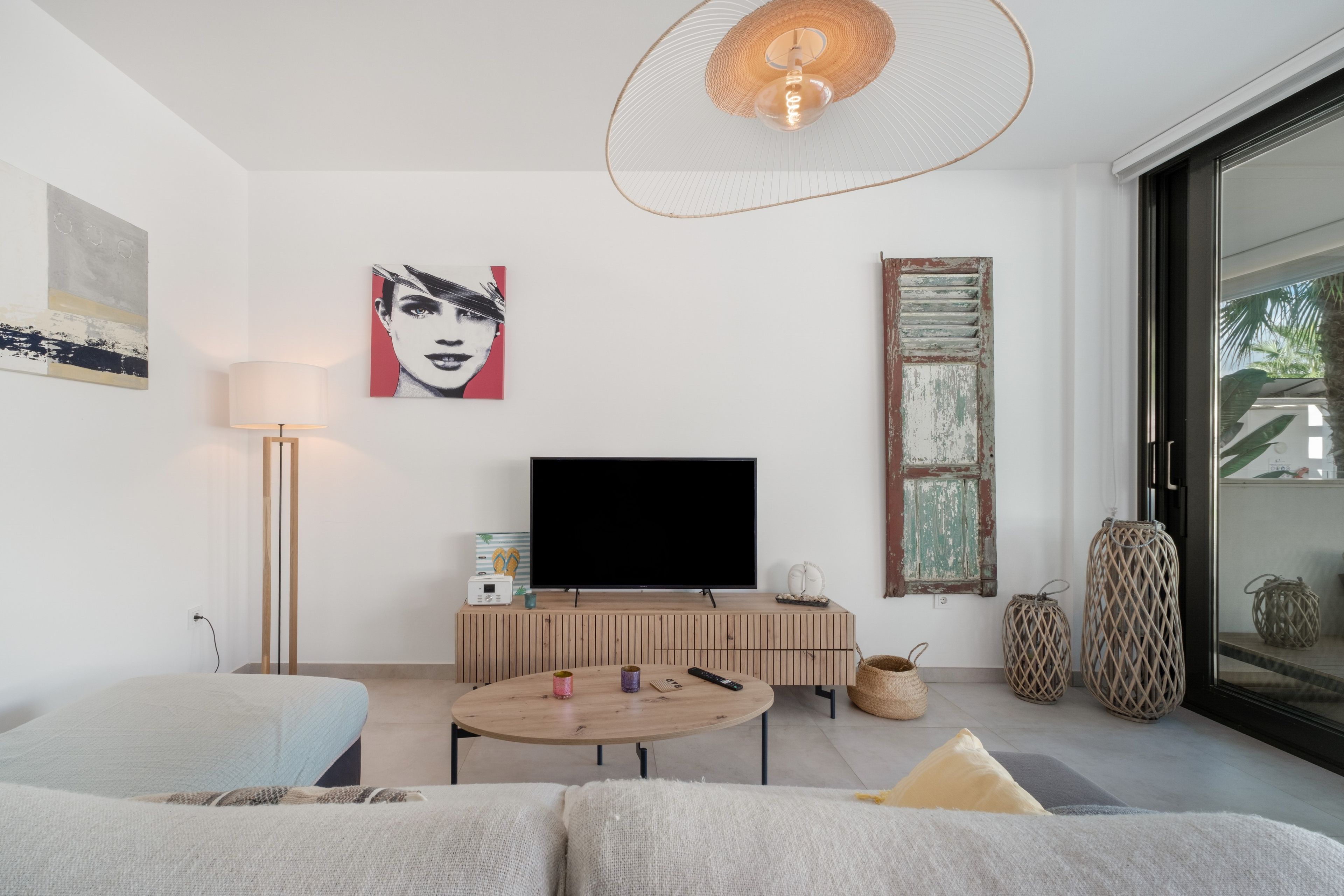 Una sala de estar moderna con un televisor de pantalla plana, una mesa de centro redonda de madera, un sofá y un ventanal con vista a palmeras.