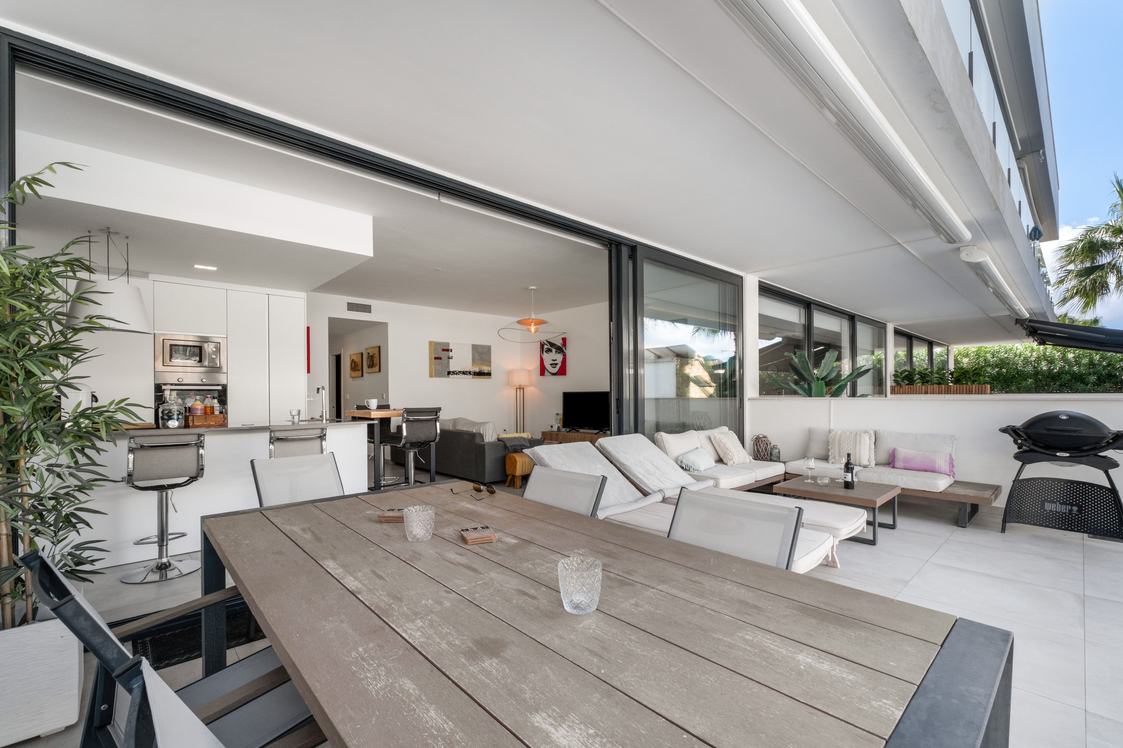 Un espacio exterior moderno con mesa de madera, sofás blancos y una parrilla.