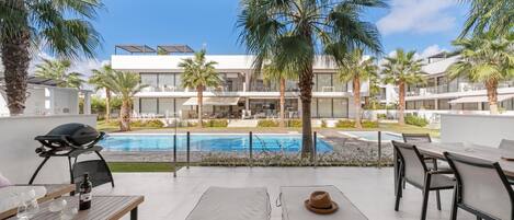 A modern outdoor lounge area with a pool, palm trees, and a view of a contemporary building.