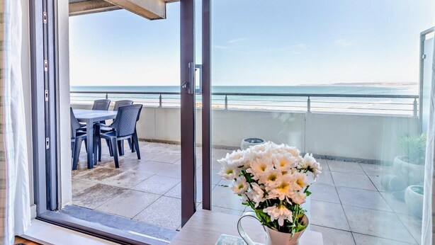 A balcony with a table and chairs, a vase of flowers, and a view of the ocean. - 3 Bed in Bideford (oc-41059) (Westward Ho!)