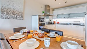 A modern kitchen with a dining area, featuring a wooden table set for breakfast, a microwave, and a stainless steel oven. - 3 Bed in Bideford (oc-41059) (Westward Ho!)