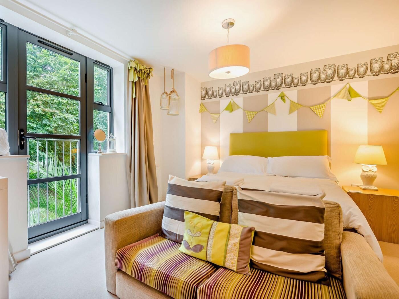 A hotel room with a bed, a sofa, a window with curtains, a wall-mounted lamp, and a decorative owl banner.