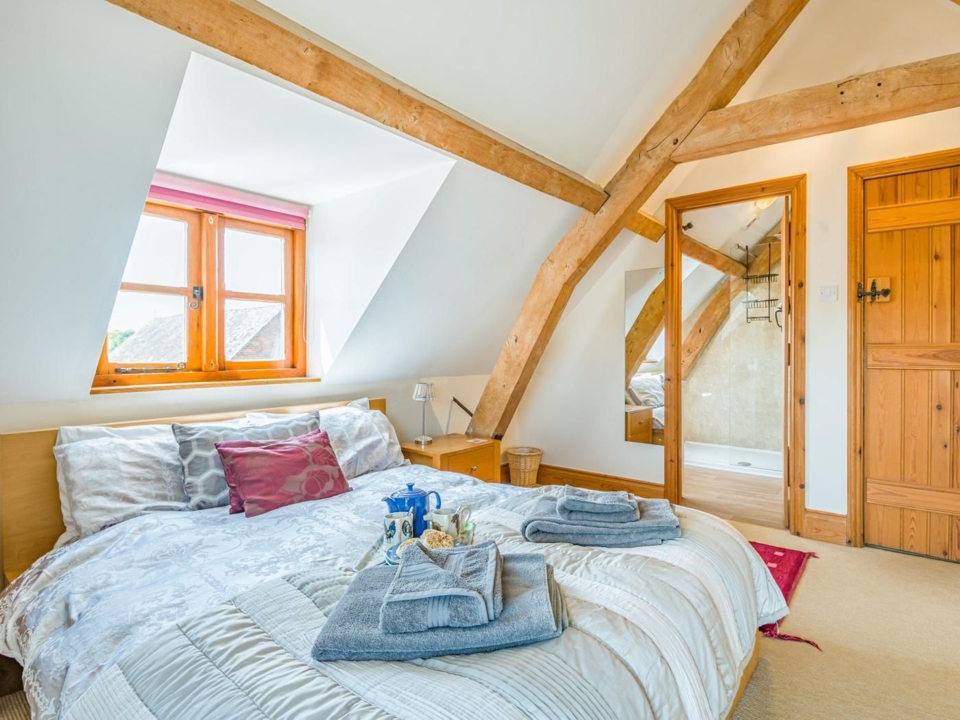 A bedroom with a sloped ceiling, a skylight, a bed with pillows and a blanket, a bedside table with a lamp, and a wooden door.