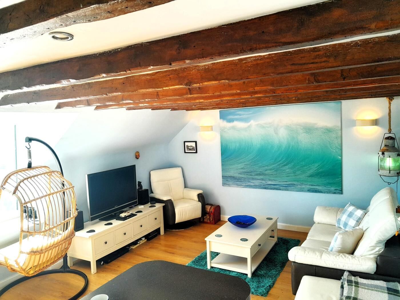 A cozy living room with a wooden ceiling, a television, a white sofa, a blue bowl on a small table, and a large wave painting on the wall.