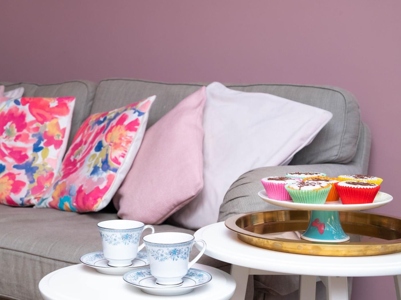 A cozy seating area with a grey sofa, colorful pillows, a small round table with a tray holding cupcakes, and a set of white cups with blue floral designs.
