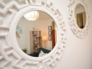 Ein Zimmer mit einem runden Spiegel, einem Bücherregal, einer Lampe und einem fensterbepflanzten Fenster.
