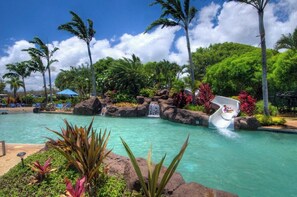 A pool with a water slide, surrounded by tropical plants and palm trees. - Stunning Ocean View Condo on the beach w AC, Kiahuna Plantation, Poipu Beach (Koloa)